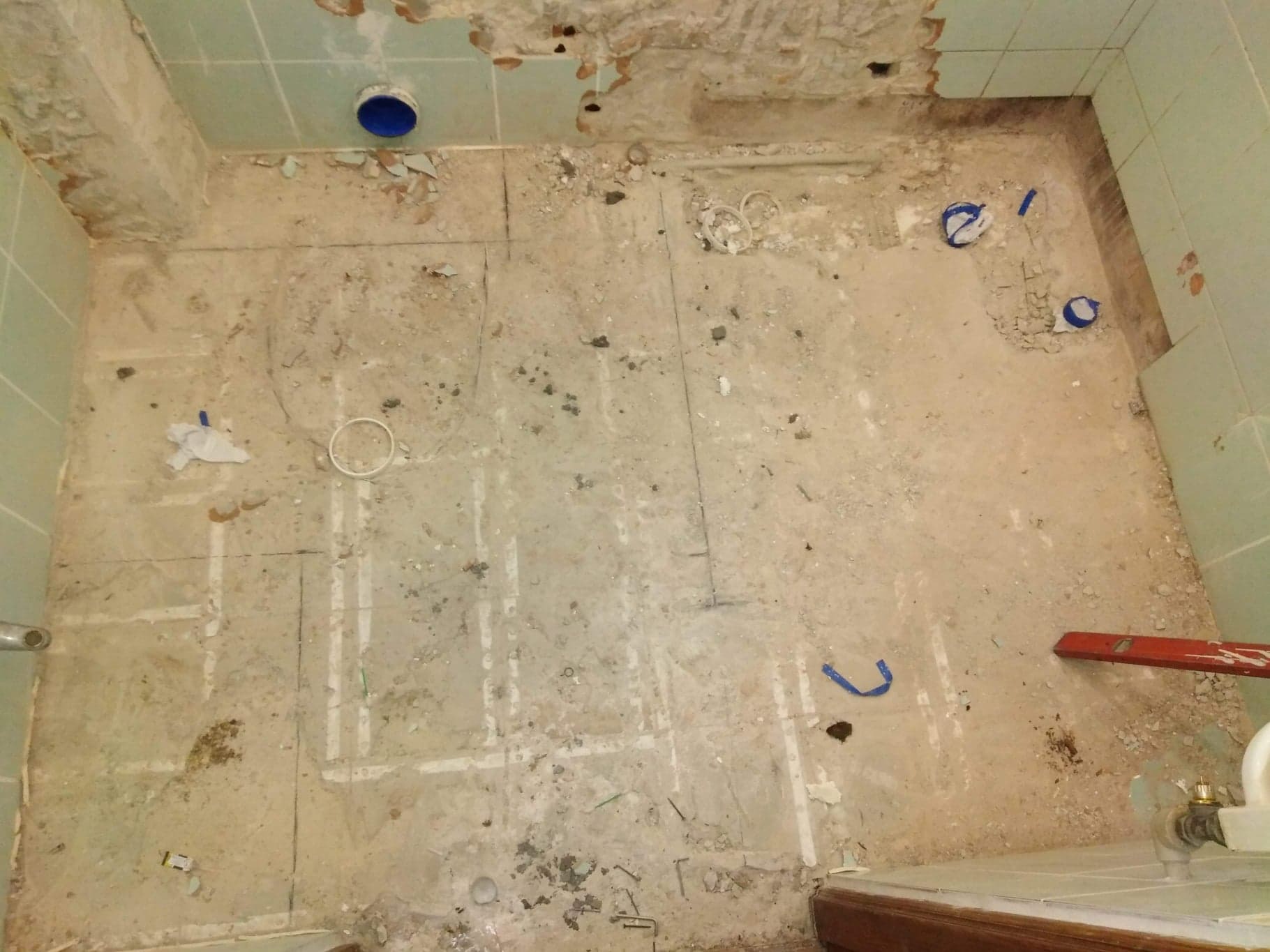 Łazienka w trakcie demontażu płytek, widoczne ślady po starych płytkach, pozostałości kleju, fragmenty instalacji i poziomica przy ścianie.