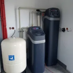 Zestaw urządzeń do filtracji i zmiękczania wody w pomieszczeniu: biały zbiornik ciśnieniowy z niebieską naklejką, dwa ciemnogranatowe filtry kolumnowe z elektronicznymi wyświetlaczami, połączone...