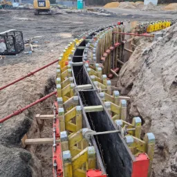 Wykonanie wykopu z szalunkiem, widoczne żółto-czerwone podpory i stalowe zbrojenie, w tle koparka CAT na placu budowy.