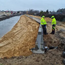 Dwóch pracowników w odblaskowych ubraniach stoi przy betonowym murze wzdłuż brzegu rzeki, w tle widoczny krajobraz z budynkami i koparką.