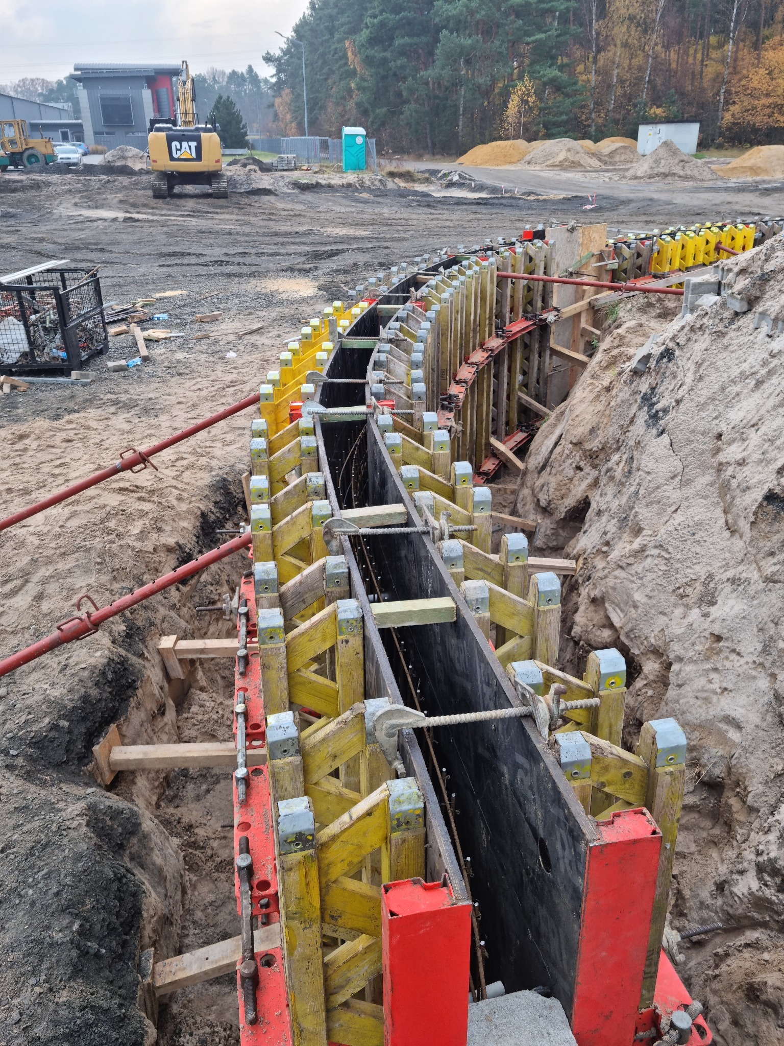 Wykonanie wykopu z szalunkiem, widoczne żółto-czerwone podpory i stalowe zbrojenie, w tle koparka CAT na placu budowy.