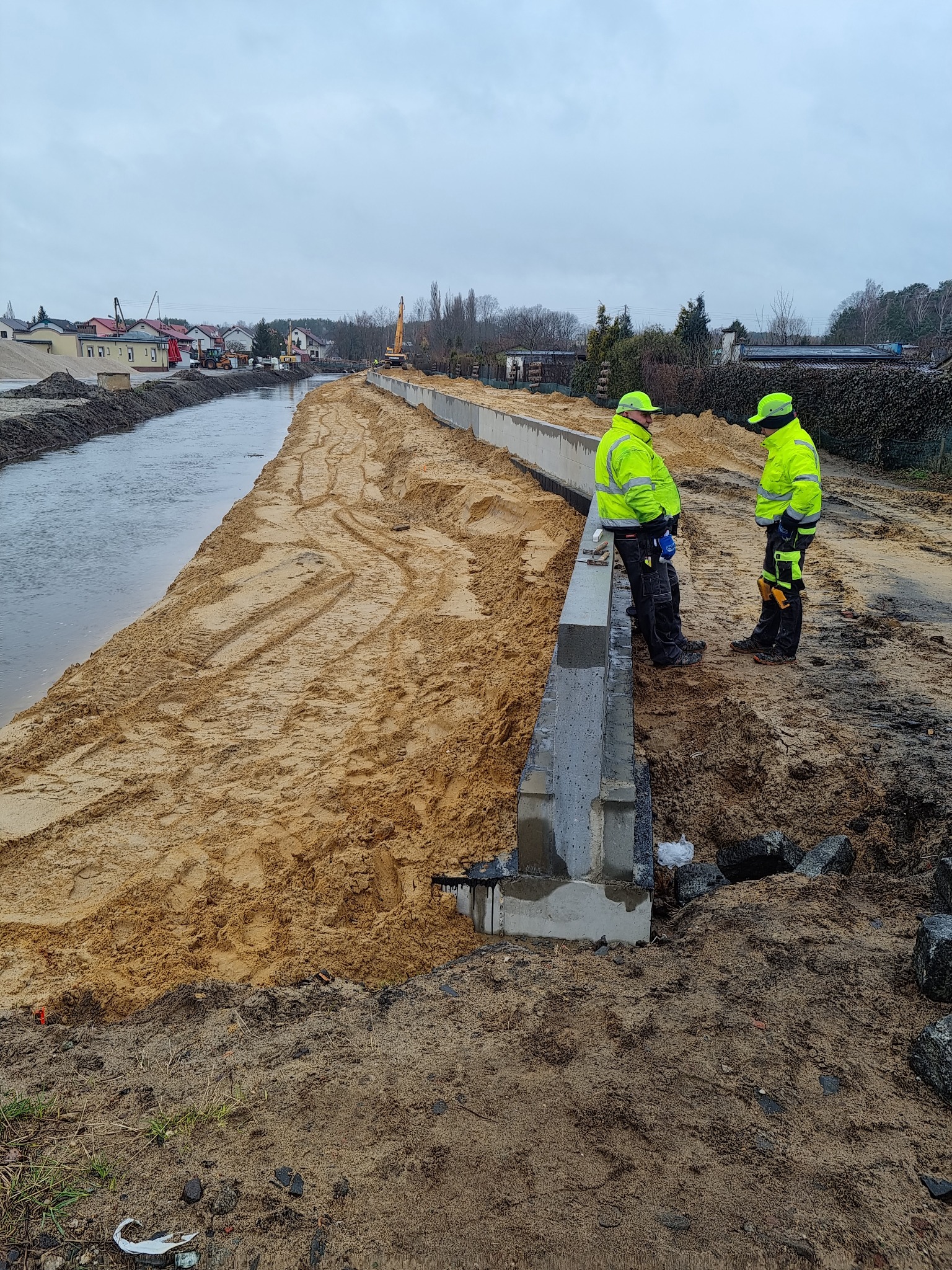 Dwóch pracowników w odblaskowych ubraniach stoi przy betonowym murze wzdłuż brzegu rzeki, w tle widoczny krajobraz z budynkami i koparką.