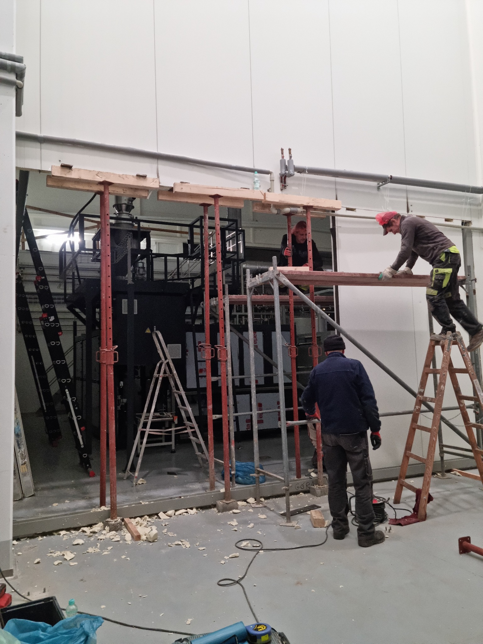 Montaż konstrukcji stalowej wewnątrz hali przemysłowej, widoczni pracownicy na rusztowaniach, fragment instalacji technologicznej w tle, ślady prac budowlanych na podłodze.
