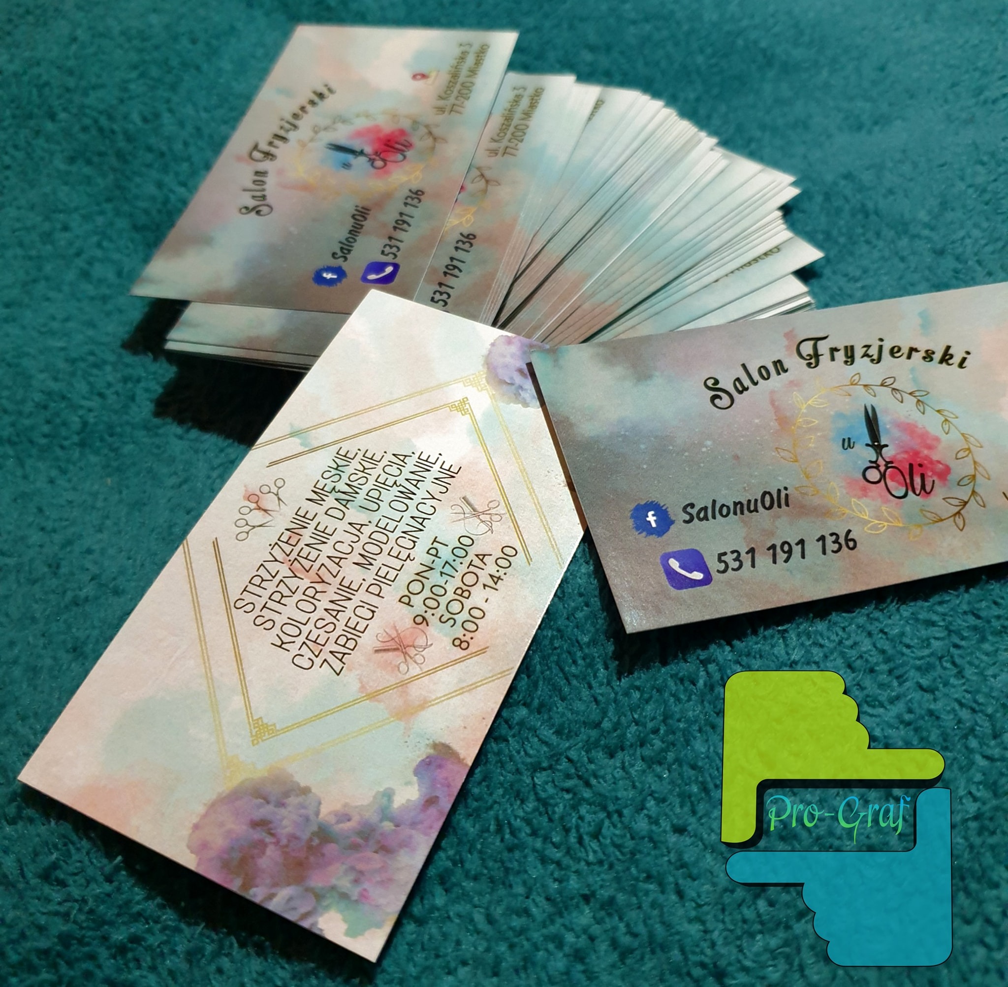 Stos wizytówek salonu fryzjerskiego z akwarelowym tłem i informacjami o usługach, numerze telefonu, adresie i godzinach otwarcia, leżących na turkusowym tle.