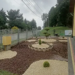 Kształtny ogród z jasnym i brązowym grysem, obrzeżami z betonowych elementów, niskimi krzewami i formowanym drzewkiem, widok z boku na posesję z metalowym ogrodzeniem.