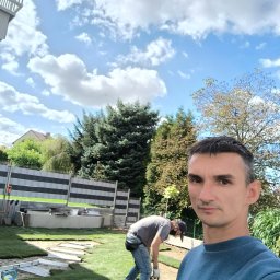 Usługi ogrodnicze - Układanie trawy z rolki w ogrodzie. Widoczny pracownik w trakcie pracy, rozłożone rolki trawy, narzędzia i fragment ogrodzenia. Słoneczny dzień, błękitne niebo z chmurami.