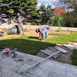 Usługi ogrodnicze - Układanie trawy z rolki w ogrodzie. Widoczne narzędzia, trawa, pracownicy i elementy dekoracyjne. Częściowo ułożona trawa i kamienne płyty chodnikowe.