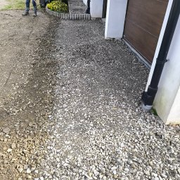 Usługi ogrodnicze - Układanie kamiennego podjazdu przy garażu. Widoczny pracownik z grabiami wyrównujący teren pod kamienie. Częściowo ułożona nawierzchnia z jasnych kamieni.