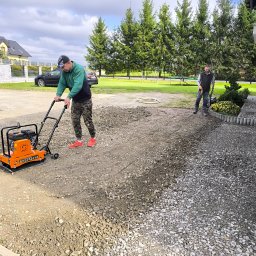 Usługi ogrodnicze - Praca zagęszczarką na żwirowym podłożu, przygotowanie terenu pod budowę. Dwóch mężczyzn w trakcie pracy, w tle dom i iglaki.