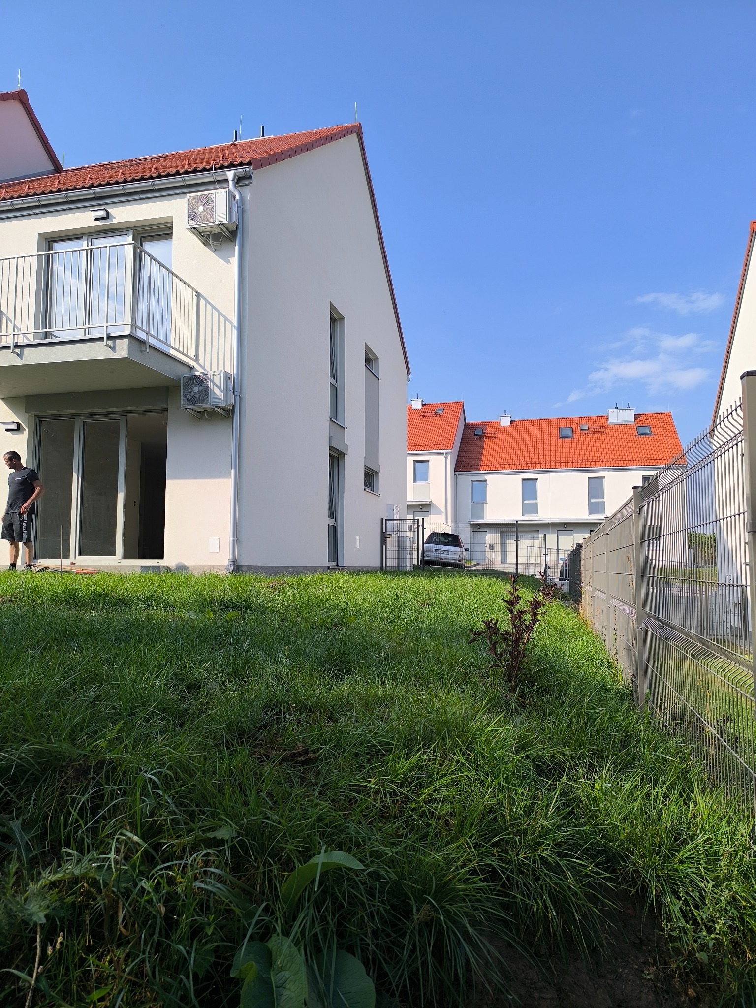 Nowoczesne szeregówki z balkonami i trawnikiem, widok z poziomu gruntu. Mężczyzna w tle przy wejściu do jednego z budynków. Metalowe ogrodzenie wzdłuż trawnika.
