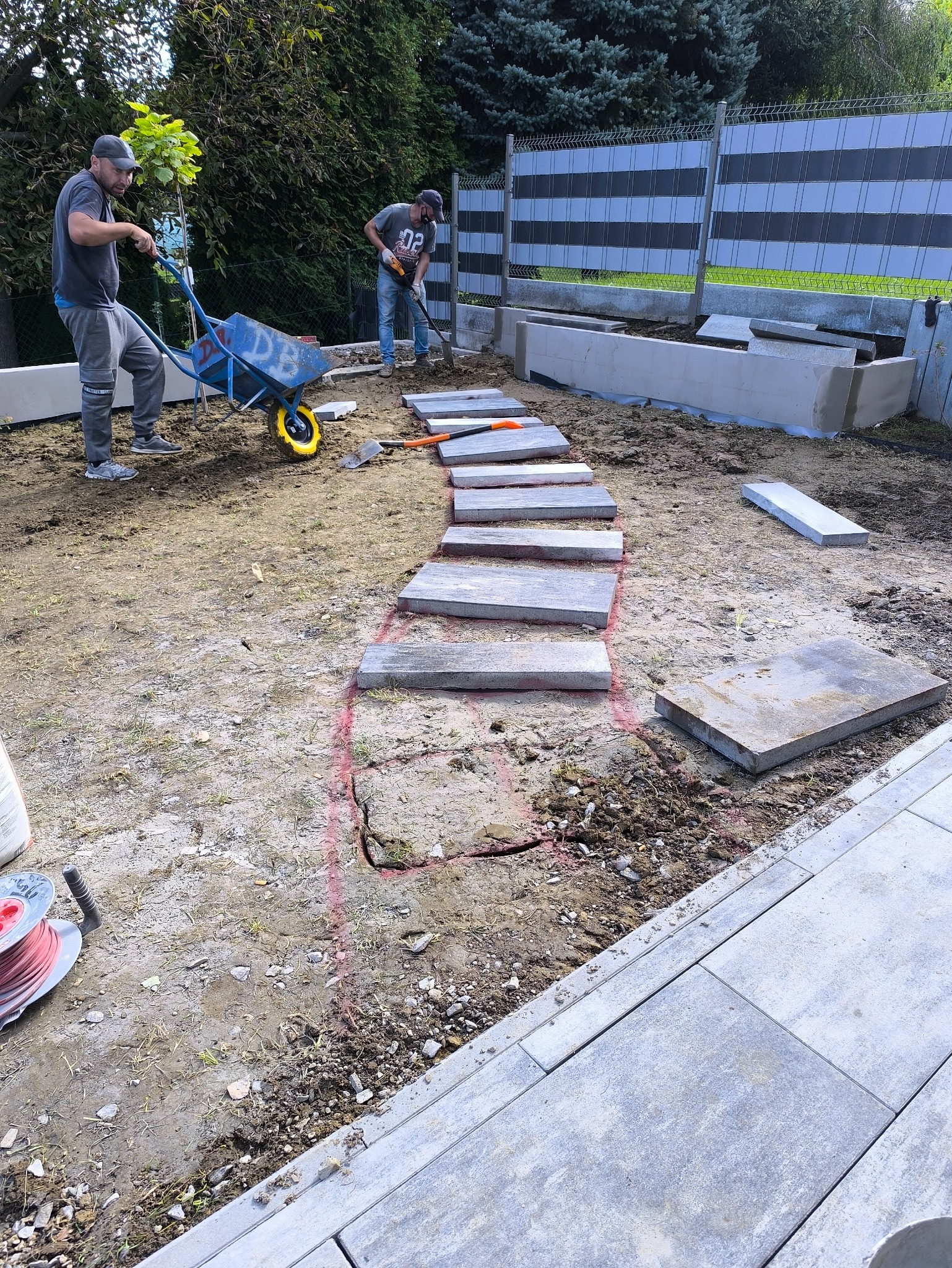 Układanie ścieżki z płyt w ogrodzie. Dwóch mężczyzn pracuje: jeden z taczką, drugi z łopatą. Widoczne narzędzia i materiały budowlane. Etap prac w słoneczny dzień.