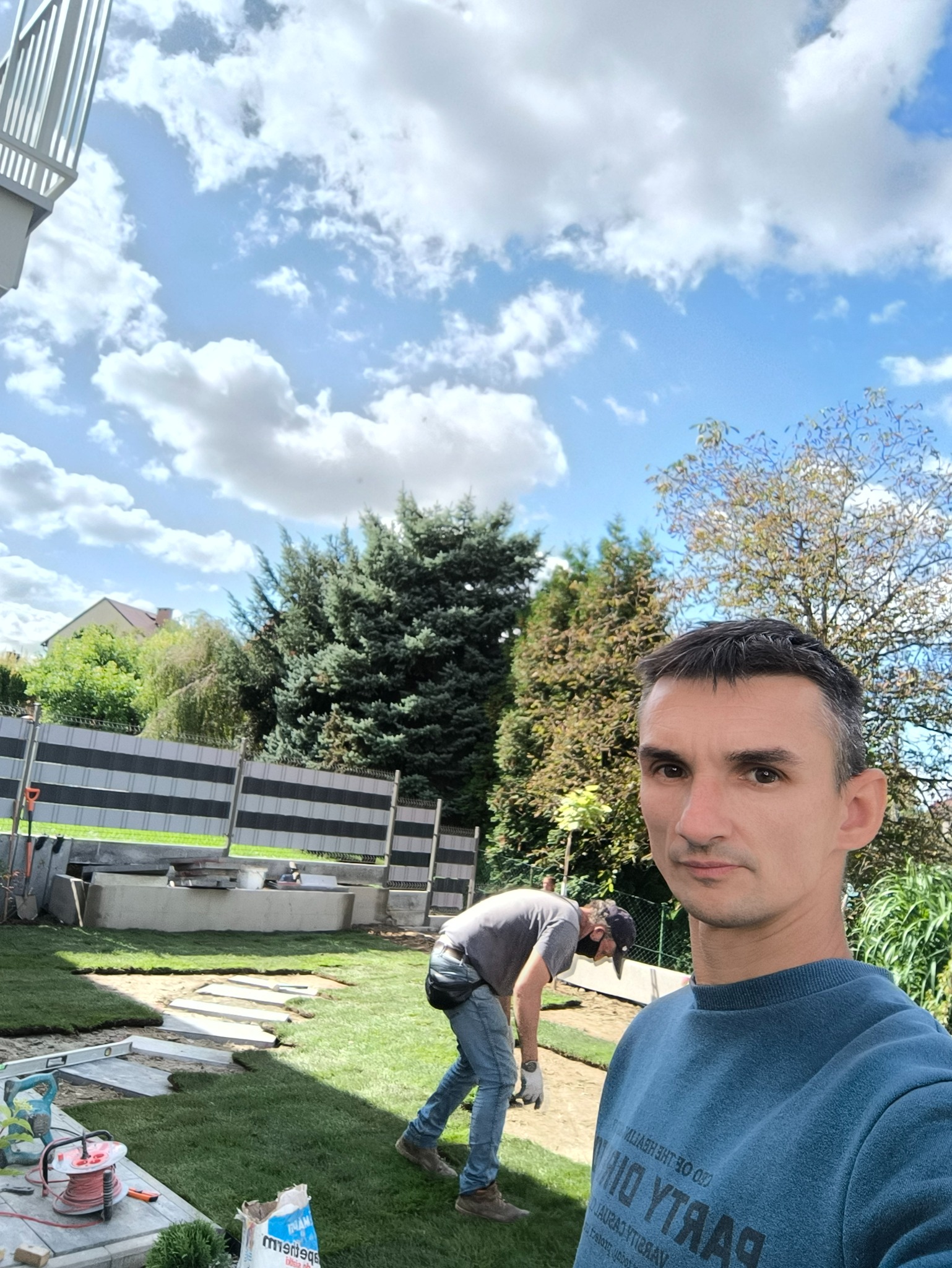 Układanie trawy z rolki w ogrodzie. Widoczny pracownik w trakcie pracy, rozłożone rolki trawy, narzędzia i fragment ogrodzenia. Słoneczny dzień, błękitne niebo z chmurami.