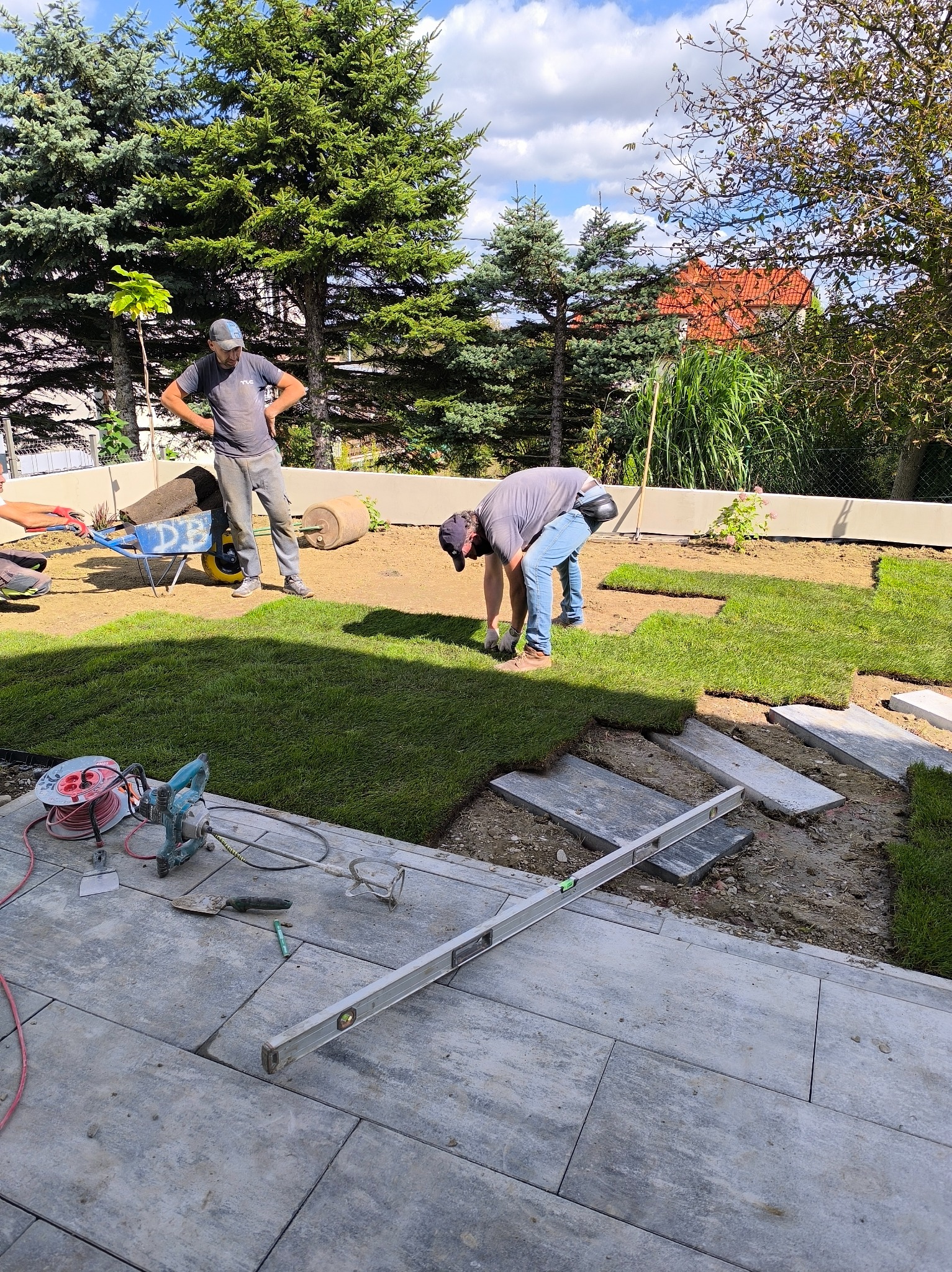 Układanie trawy z rolki w ogrodzie. Widoczne narzędzia, trawa, pracownicy i elementy dekoracyjne. Częściowo ułożona trawa i kamienne płyty chodnikowe.