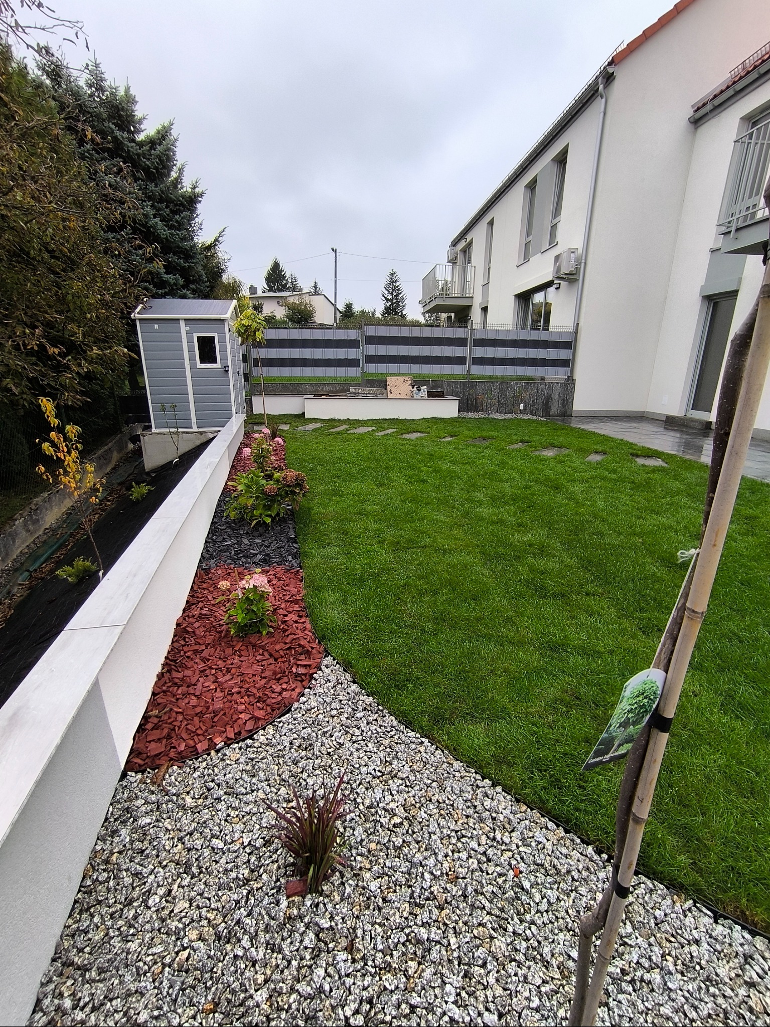 Nowoczesny ogród z równo przystrzyżonym trawnikiem, rabatami z kamieni, kory i roślin, oraz szarą drewutnią w tle, na tle białego domu z balkonami.