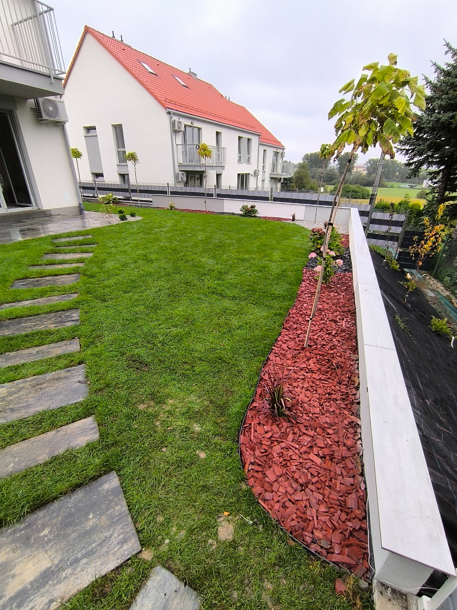 Zadbany ogród z równo przystrzyżonym trawnikiem, ścieżką z kamiennych płyt i rabatą z czerwoną korą. W tle nowoczesny dom z balkonami i zielenią.