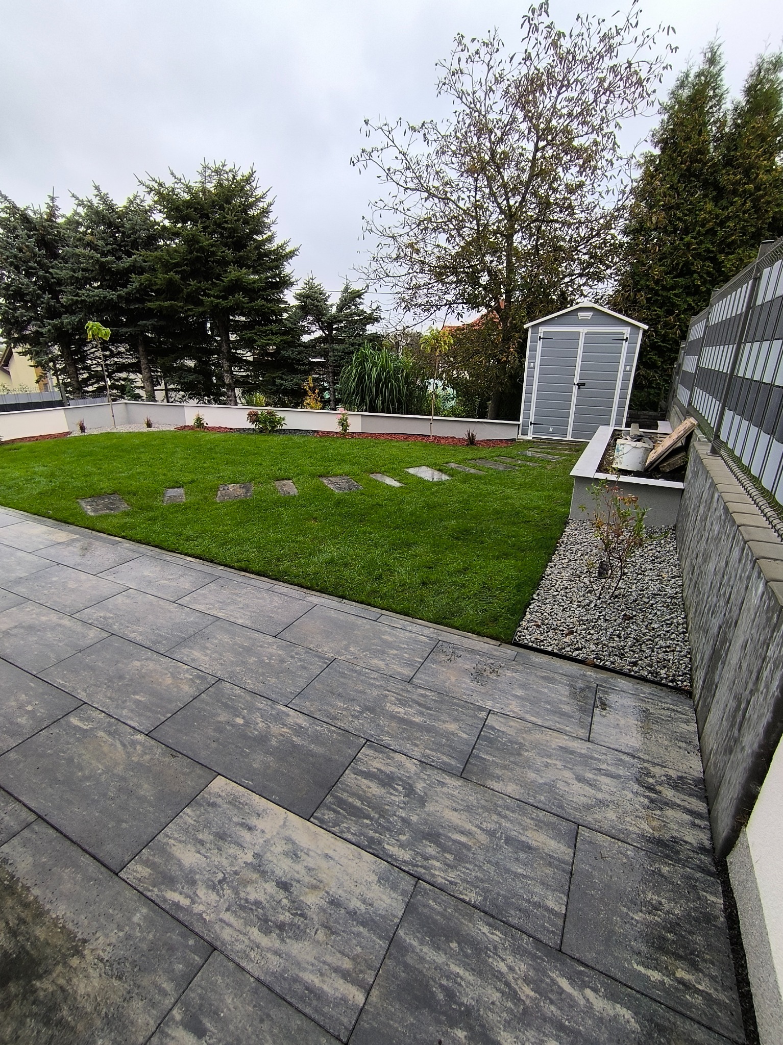 Nowoczesny ogród z równo przystrzyżonym trawnikiem, ścieżką z szarych płyt, ozdobnym kamieniem i szarą szopą narzędziową w tle, w pochmurny dzień.