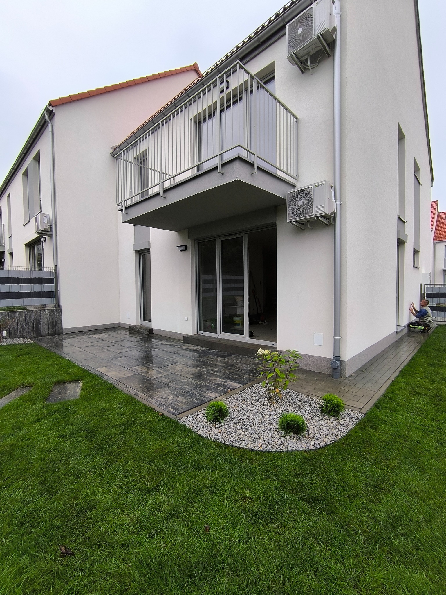 Nowoczesny dom z balkonem i tarasem wyłożonym kamienną kostką. Uwagę przyciąga rabata z krzewami i kamieniami. Osoba przy ścianie, prawdopodobnie wykonująca prace wykończeniowe.