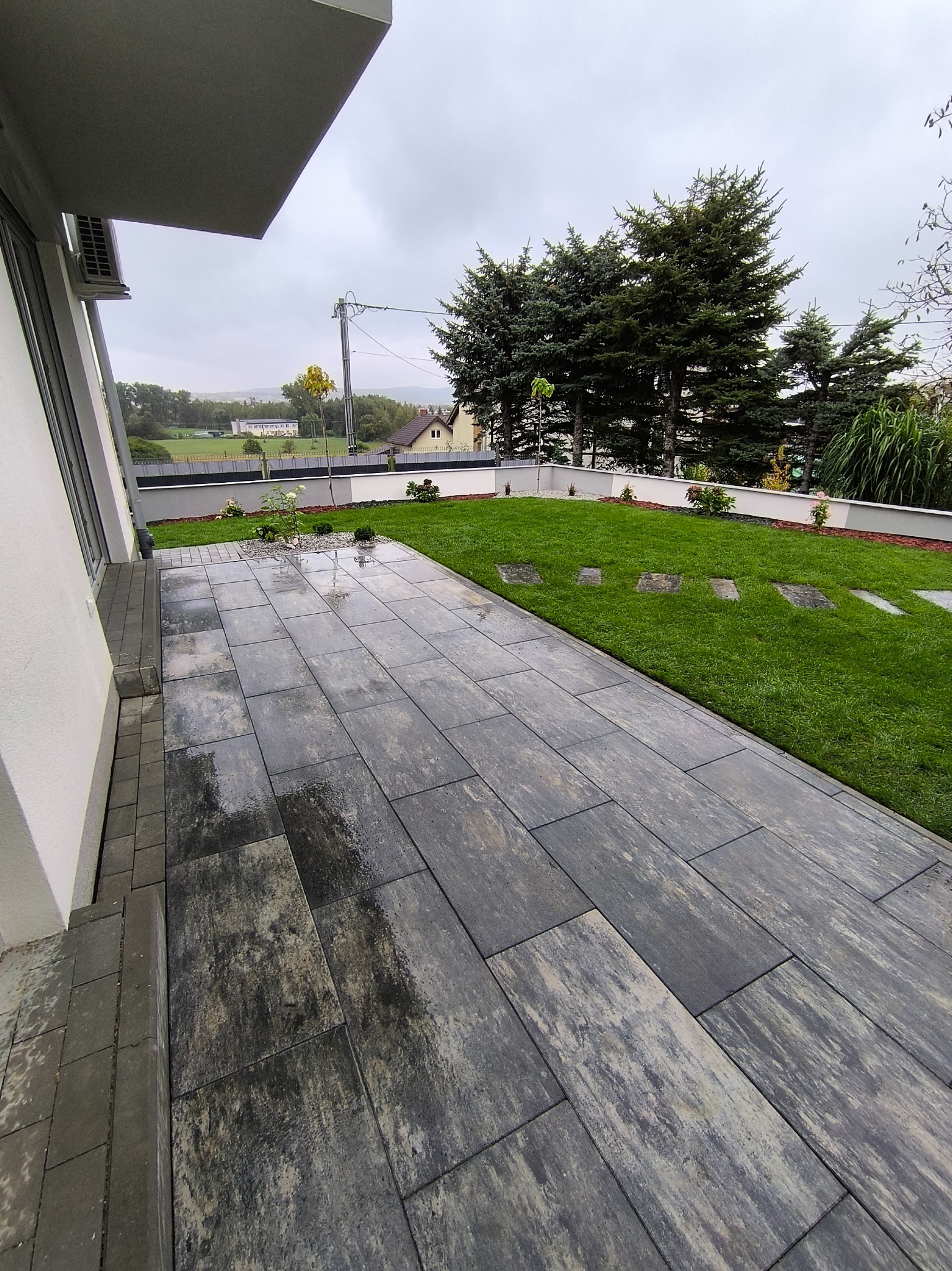Nowoczesny taras z szarych płyt betonowych, przylegający do budynku. Widoczny trawnik z elementami dekoracyjnymi i drzewa w tle. Całość utrzymana w stonowanej kolorystyce.