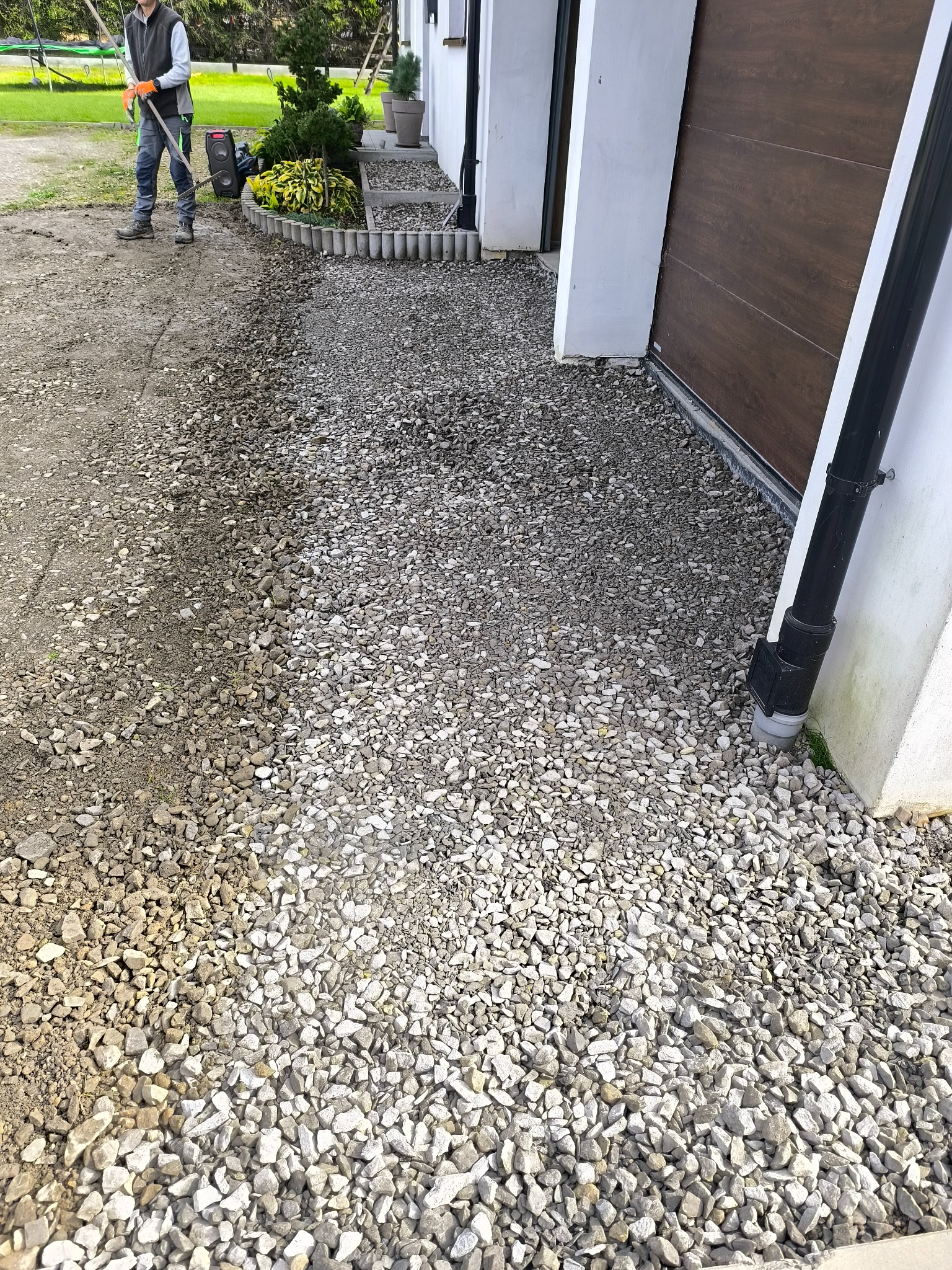 Układanie kamiennego podjazdu przy garażu. Widoczny pracownik z grabiami wyrównujący teren pod kamienie. Częściowo ułożona nawierzchnia z jasnych kamieni.