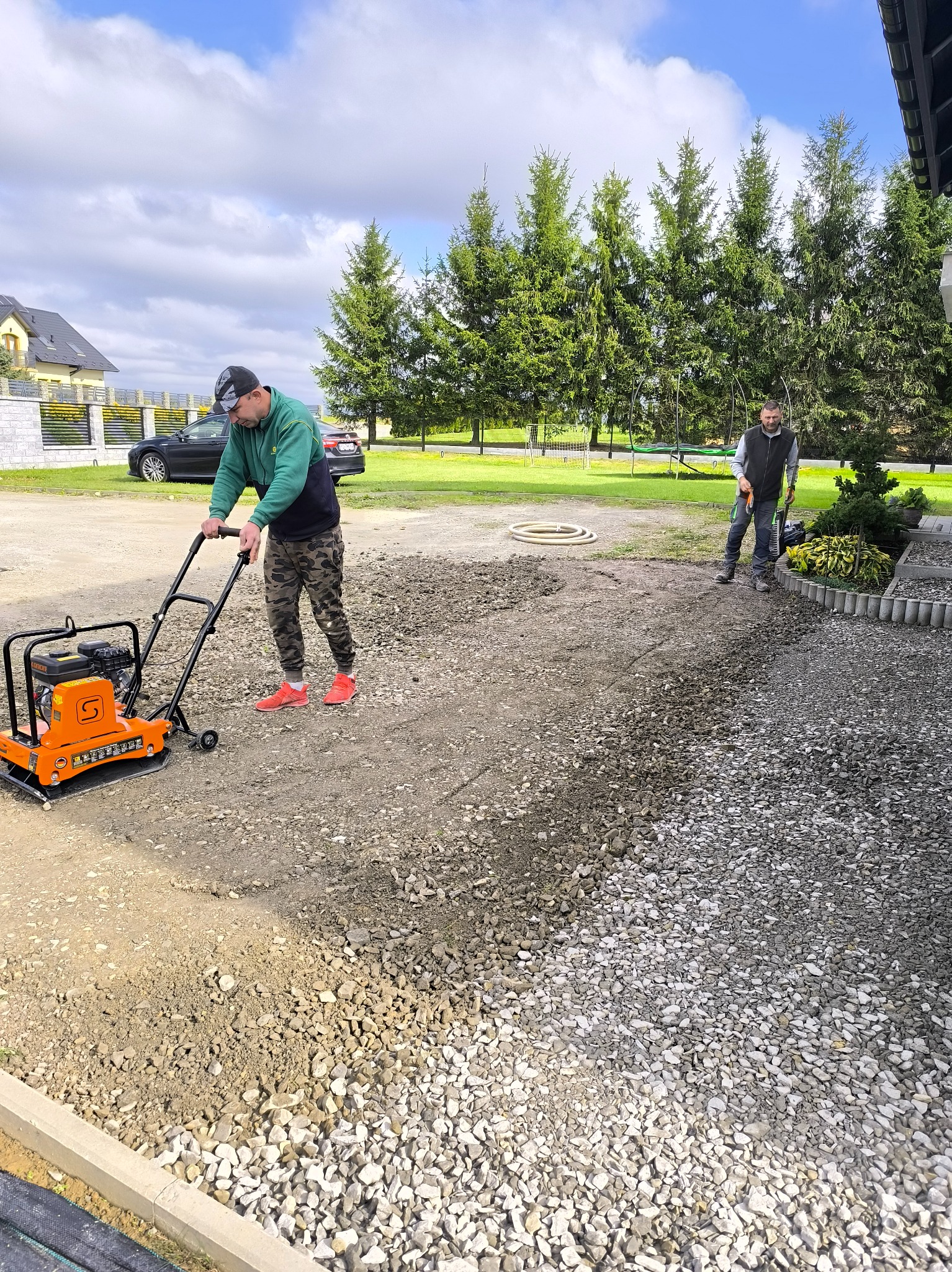 Praca zagęszczarką na żwirowym podłożu, przygotowanie terenu pod budowę. Dwóch mężczyzn w trakcie pracy, w tle dom i iglaki.