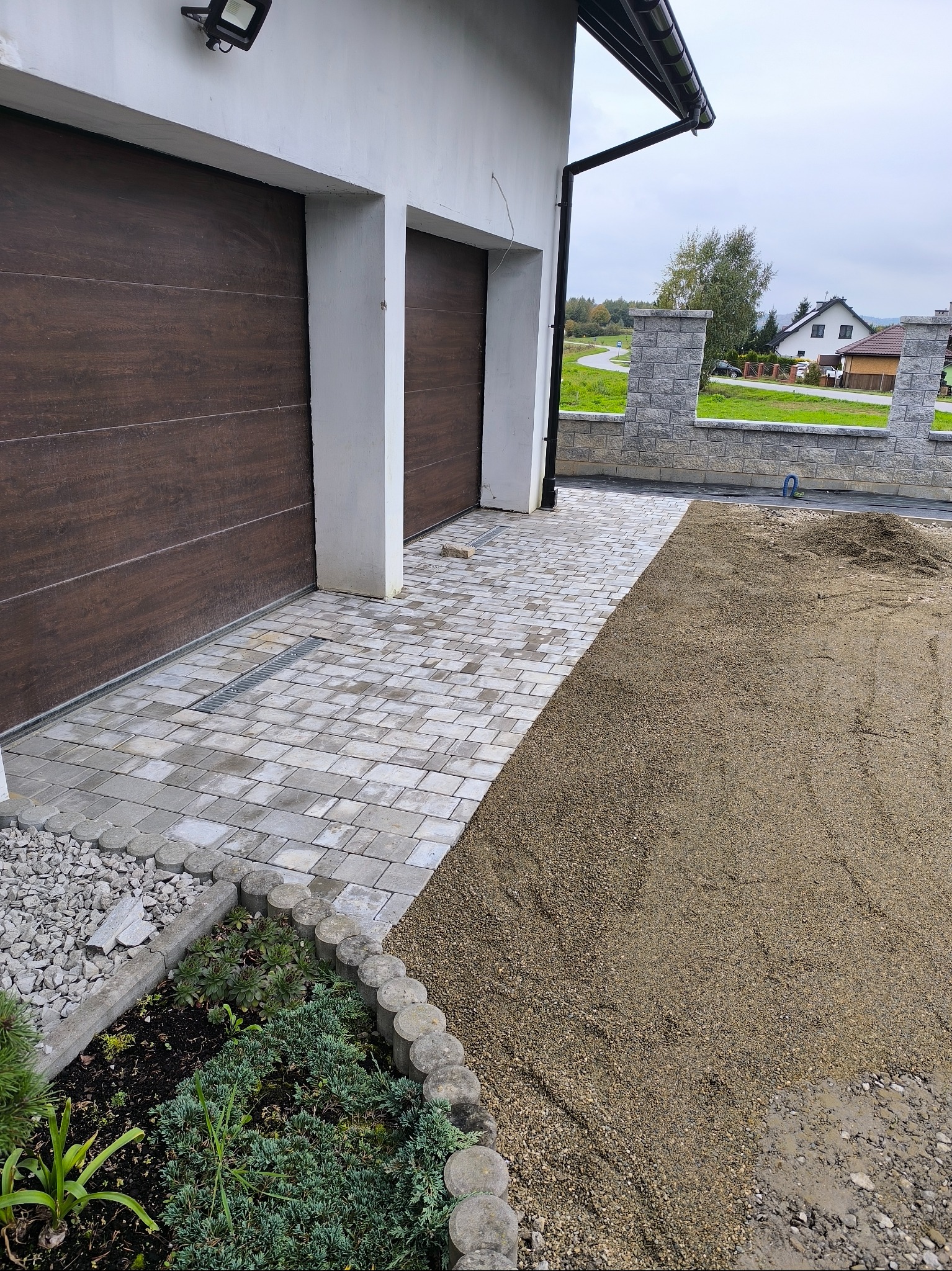 Ułożona kostka brukowa przed garażem z odpływem liniowym, obrzeża z roślinnością i przygotowany teren pod dalsze prace. Widok na dom i ogrodzenie.