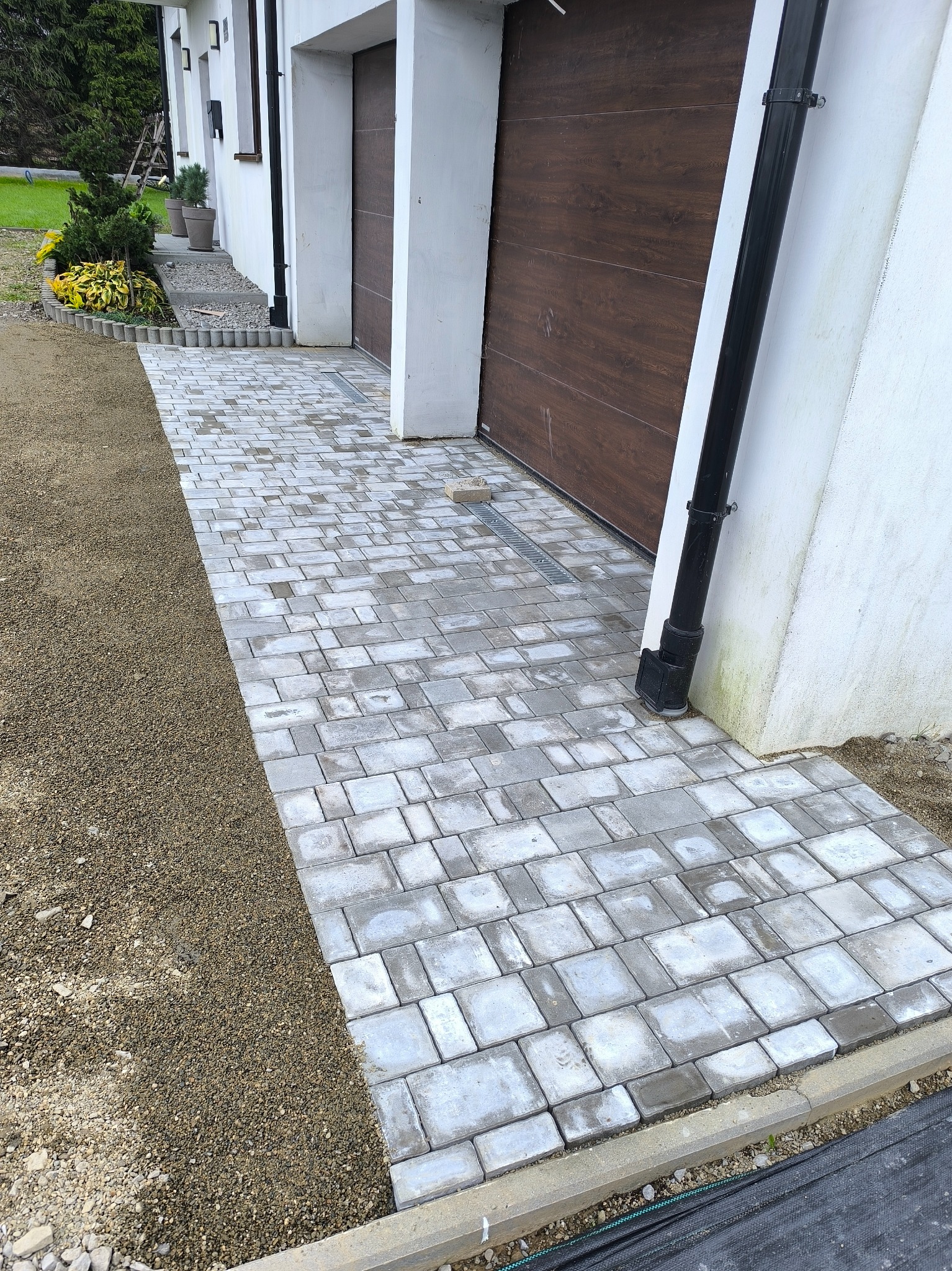 Ułożona kostka brukowa tworząca ścieżkę wzdłuż domu z brązową bramą garażową. Widoczny fragment rabaty z roślinami i krawężnik oddzielający kostkę od gruntu.