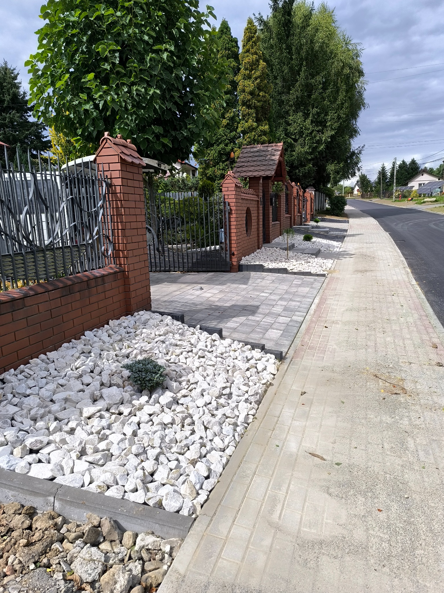 Ogród frontowy z dekoracyjnym białym kamieniem, niską roślinnością i brukowanym chodnikiem. W tle ogrodzenie z cegły i metaloplastyki.