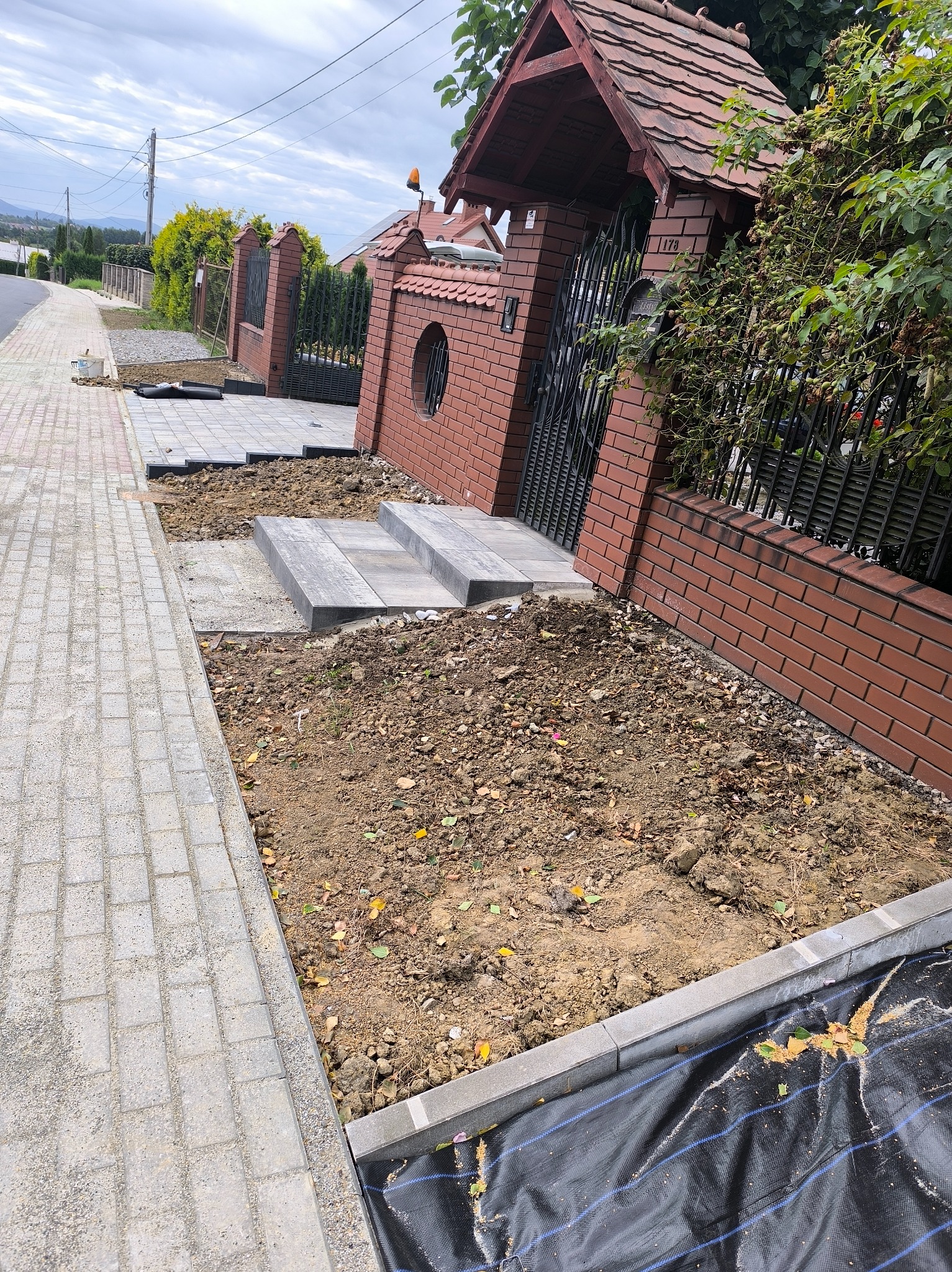 Układanie kostki brukowej przy ogrodzeniu z cegły. Widoczne schody z kostki i przygotowany teren pod dalsze prace. Fragment chodnika z kostki brukowej.