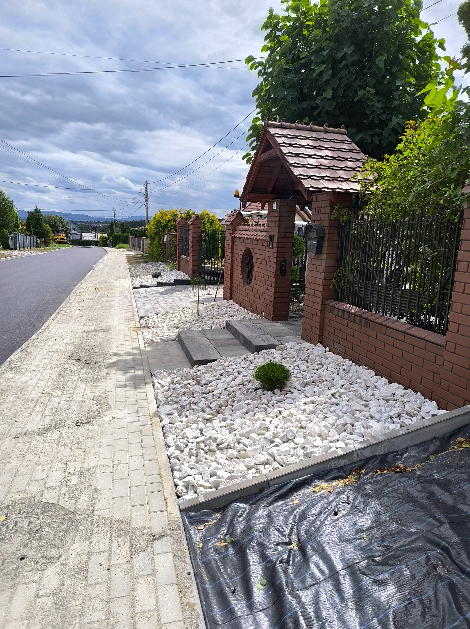 Ogród frontowy z białymi kamieniami, niską roślinnością i ceglanym ogrodzeniem z metalowymi elementami. Widoczne schody z płyt i czarna agrowłóknina.