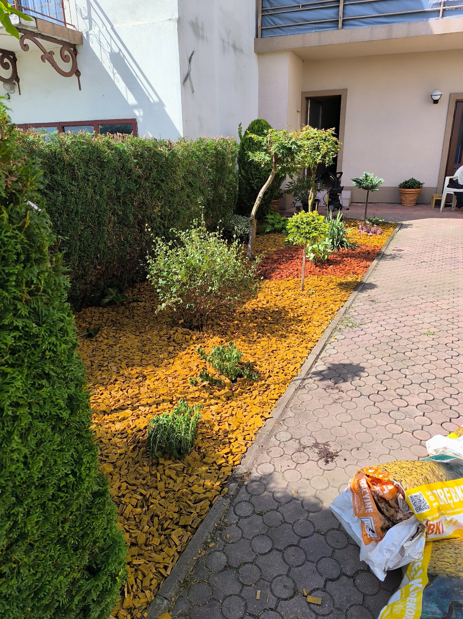 Aranżacja ogrodu w Gorlicach: rabata wysypana korą, obok chodnik z kostki. W tle żywopłot i budynek. Worki z korą leżą na chodniku.