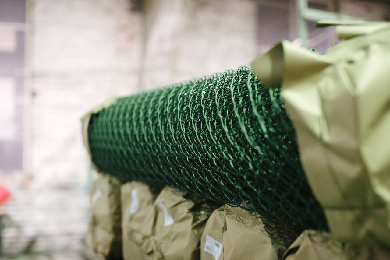 Rolka zielonej, powlekanej siatki ogrodzeniowej na palecie, zabezpieczona folią ochronną, w magazynie.