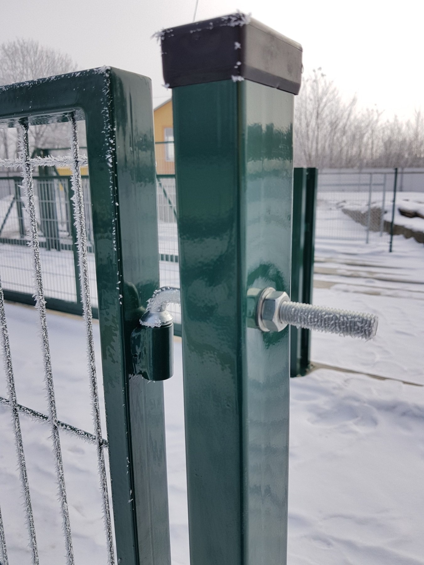 Fragment zielonej bramy z siatki ogrodzeniowej, pokrytej szronem w zimowy dzień, widoczne detale mocowania i zamek.