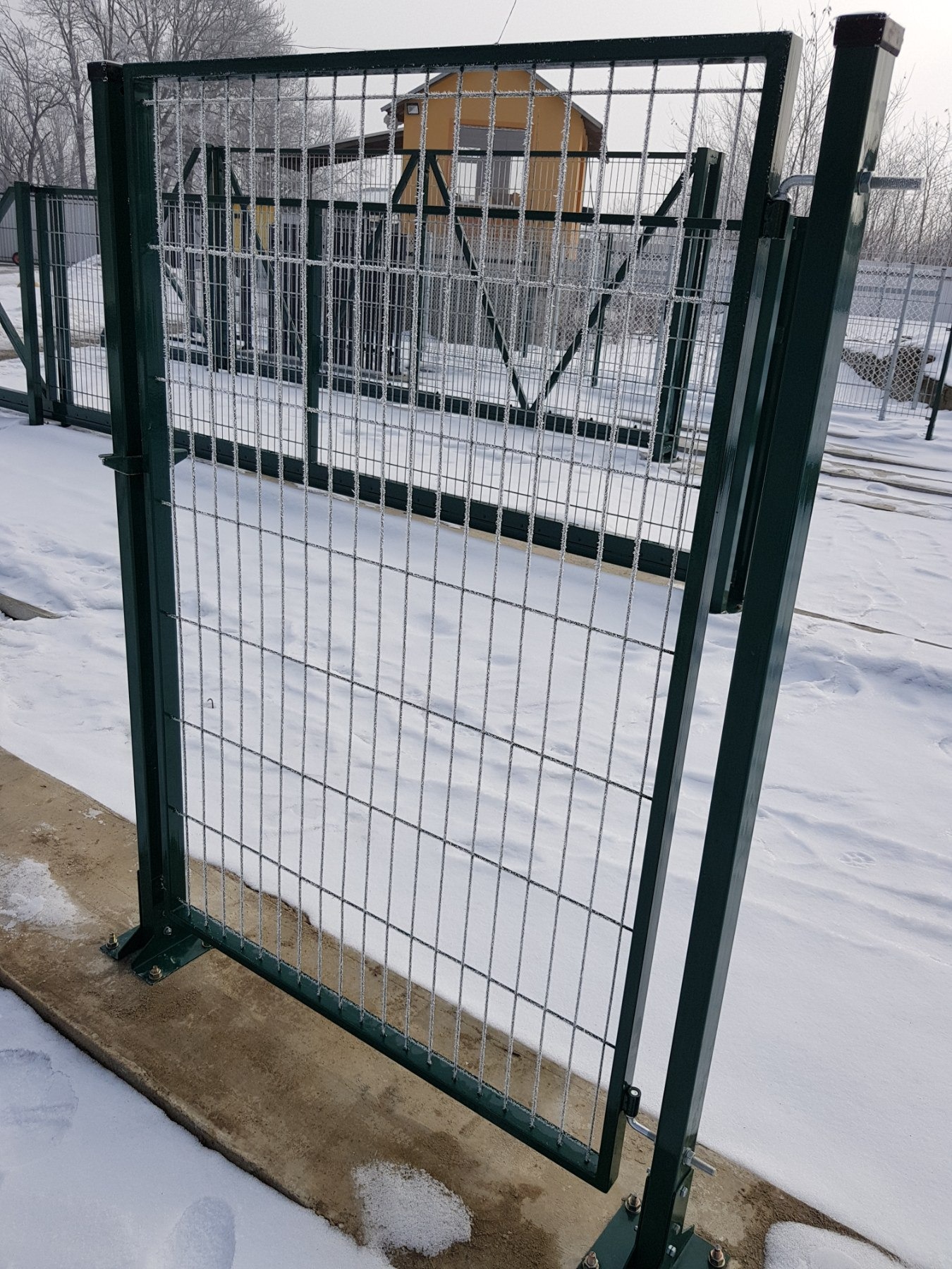Zielona, metalowa brama z siatką ogrodzeniową pokryta szronem, zamontowana na betonowej podstawie w zimowym krajobrazie.