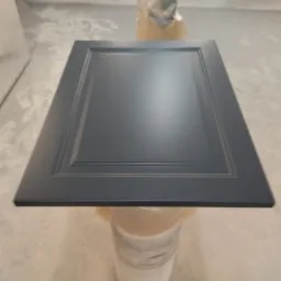 Pomalowane na szaro, frezowane drzwiczki szafki kuchennej, ustawione na statywie w pomieszczeniu lakierniczym.