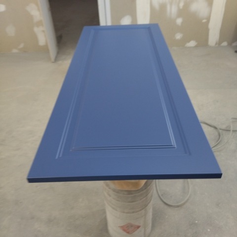 Świeżo pomalowane na niebiesko drzwiczki szafki z frezowanymi krawędziami, ustawione na wiadrze w pomieszczeniu w trakcie remontu.