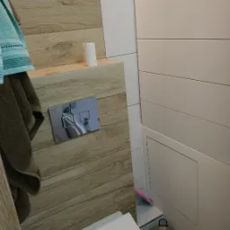 Nowoczesna toaleta podwieszana z drewnopodobną obudową, chromowanym przyciskiem spłukiwania i szczotką do WC; widoczny kosz na śmieci i ręczniki.