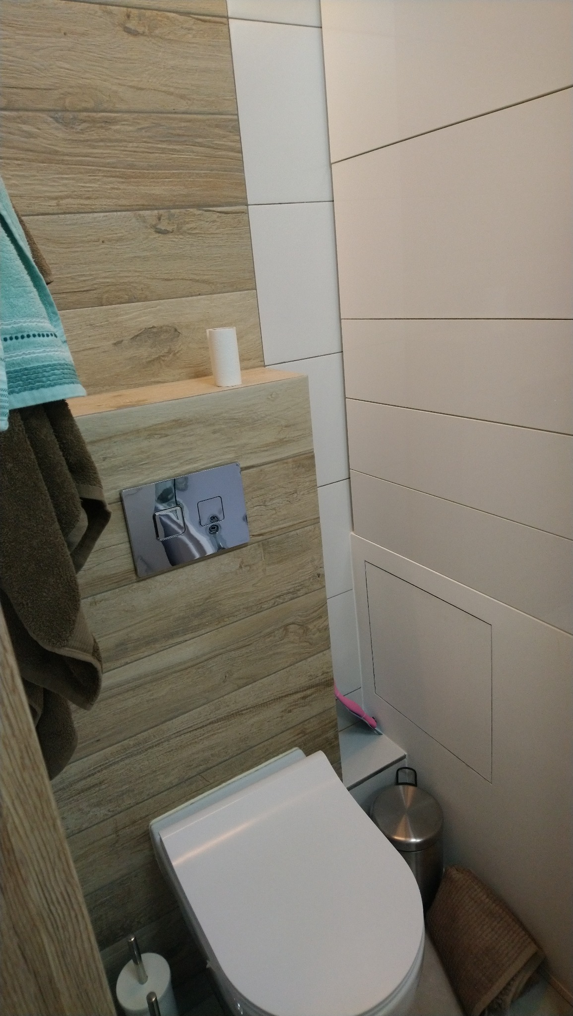 Nowoczesna toaleta podwieszana z drewnopodobną obudową, chromowanym przyciskiem spłukiwania i szczotką do WC; widoczny kosz na śmieci i ręczniki.