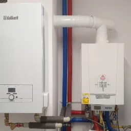 Dwa białe piece gazowe różnych marek (Vaillant i De Dietrich) zamontowane na ścianie z widocznymi rurami instalacji CO, rury izolowane termicznie, niebieski zbiornik wyrównawczy.