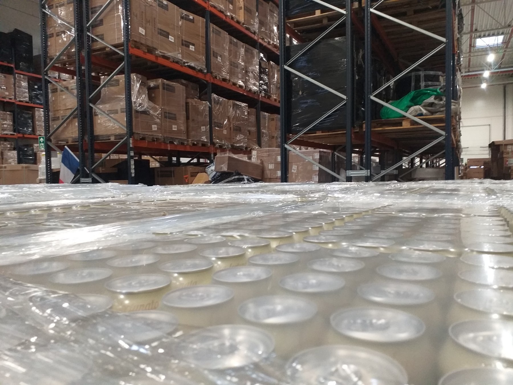 Paleta puszek z konserwami, owinięta folią, na tle regałów magazynowych z kartonami i flagą Francji w tle.