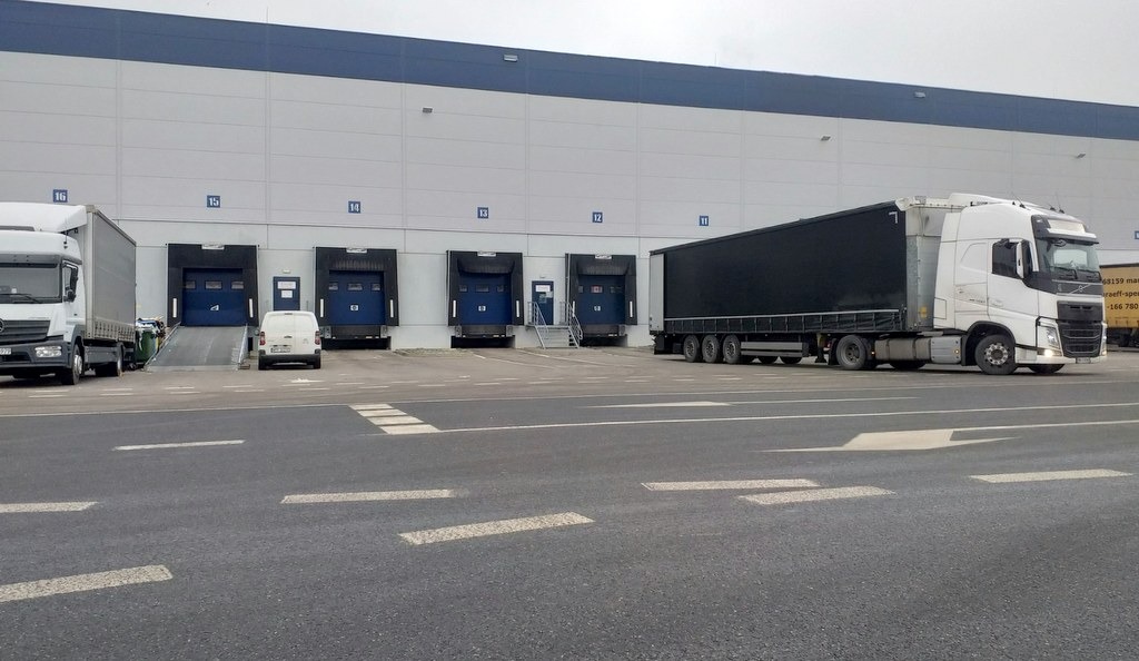 Ciężarówki stojące przed rampami załadunkowymi magazynu logistycznego, widoczny biały bus dostawczy i oznakowanie poziome.