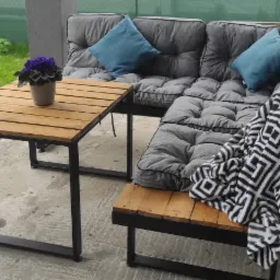 Zestaw mebli ogrodowych z metalową ramą i drewnianymi elementami: stół z kwiatami w doniczce i narożna sofa z szarymi poduchami i niebieskimi poduszkami, przykryta czarno-białym kocem...