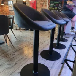 Szereg nowoczesnych, szarych krzeseł barowych z czarnymi, metalowymi nogami na tle ceglanej ściany w lokalu gastronomicznym. Widoczna tablica z menu napojów.