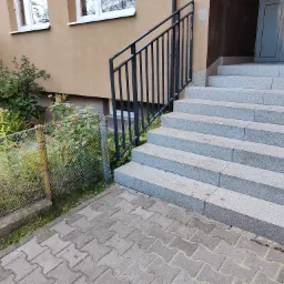 Zewnętrzne schody z szarego granitu prowadzące do wejścia budynku, z czarną, metalową balustradą po lewej stronie, widoczne z poziomu chodnika z kostki brukowej.