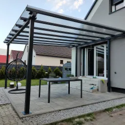 Czarna metalowa pergola z przezroczystym zadaszeniem poliwęglanowym, przylegająca do domu, z widocznym stołem ogrodowym i wiszącym fotelem na tle zadbanego ogrodu.