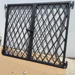 Dwu-skrzydłowa, czarna, metalowa brama z romboidalną kratą, leżąca na kartonie przed garażem, z dekoracyjnymi zawiasami i zamkiem.