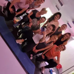 Grupa uśmiechniętych kobiet w sportowych strojach robi sobie selfie w lustrze w sali fitness, widoczna mata do ćwiczeń i hantle w tle.
