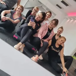 Grupa uśmiechniętych kobiet w strojach sportowych pozuje do selfie w lustrze na tle sali gimnastycznej, widać stepy i maty podłogowe.