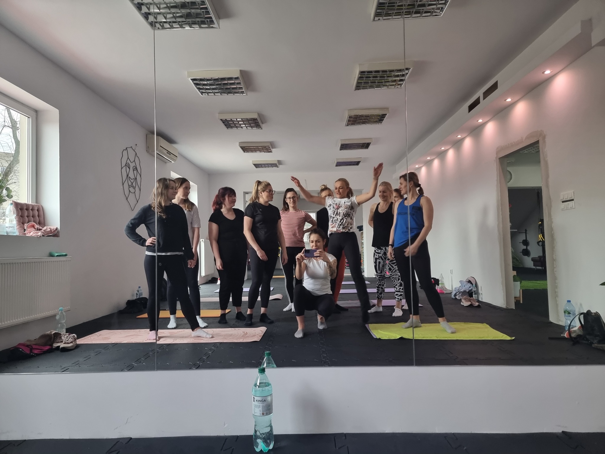 Grupa kobiet w sali, niektóre na matach do ćwiczeń, pozuje do zdjęcia robionego telefonem przez jedną z nich, widok odbity w lustrze.