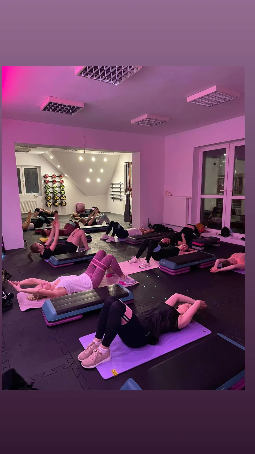 Grupa kobiet ćwiczy na matach i stepach w sali fitness oświetlonej różowym światłem, wykonując ćwiczenia wzmacniające mięśnie brzucha i pośladków.