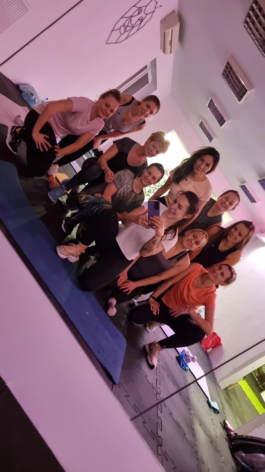Grupa uśmiechniętych kobiet w sportowych strojach robi sobie selfie w lustrze w sali fitness, widoczna mata do ćwiczeń i hantle w tle.